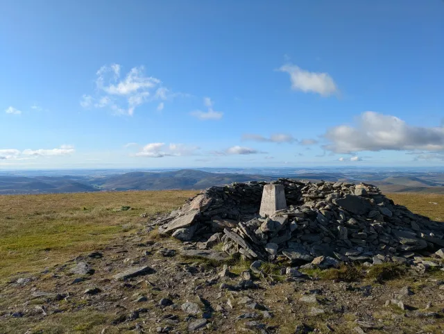 A trig point and cairn on a mountain ridge above a wide landscape under blue sky