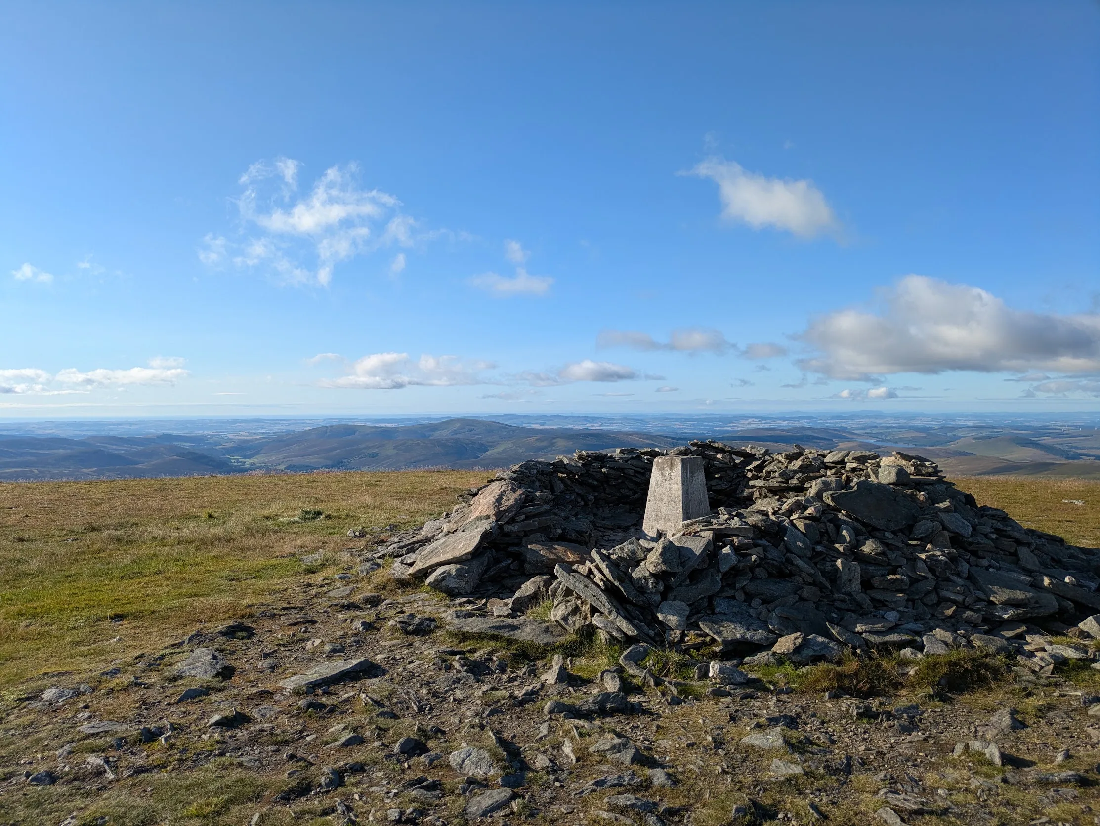 A trig point and cairn on a mountain ridge above a wide landscape under blue sky