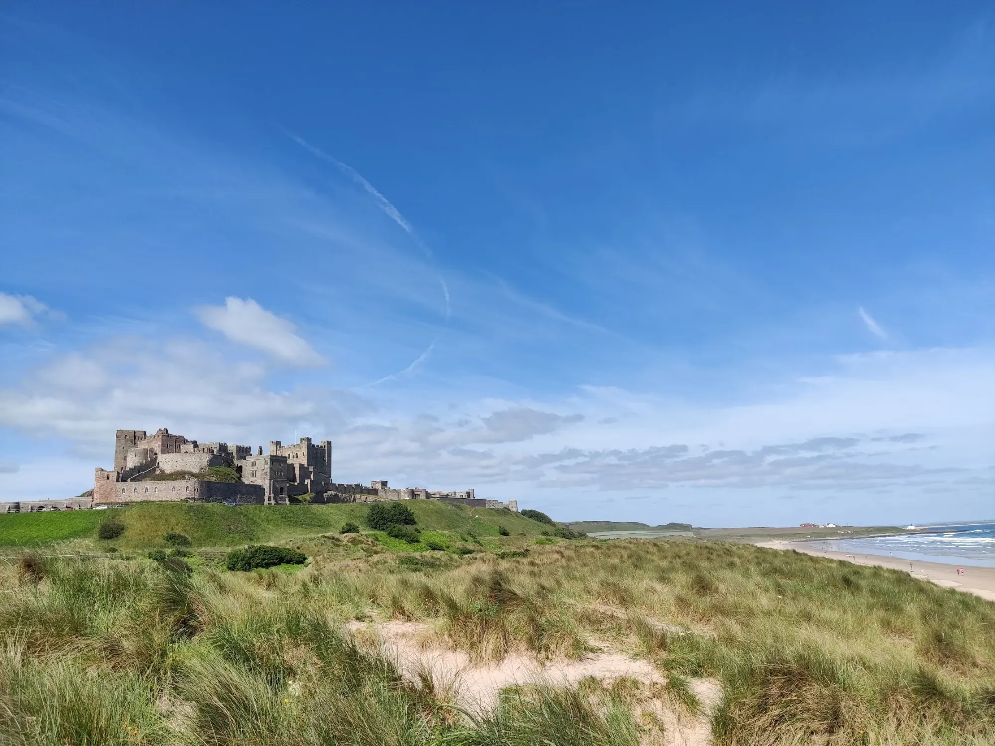 A stone castle above a sandy beach and grassy dunes beneath a blue sky