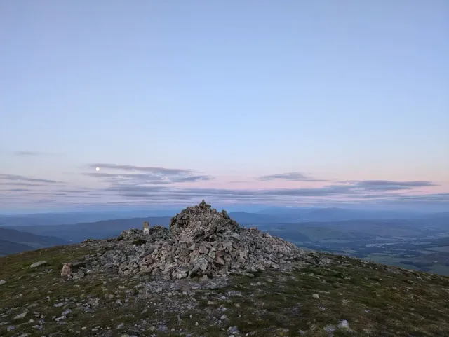 A rocky summit cairn beneath a pink-blue dawn sky, with a pale moon over distant hills