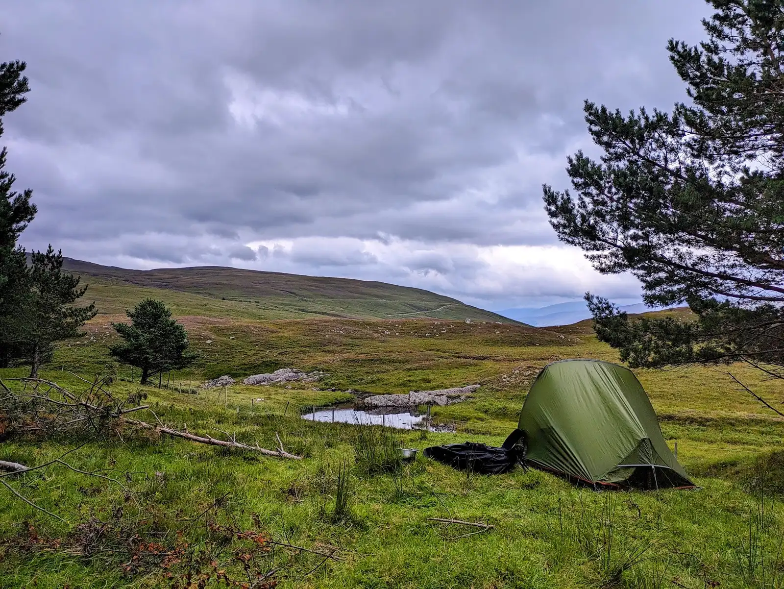 A green backpacking tent pitched by a small lochan on Scottish moorland, pack and pot sat on the grass