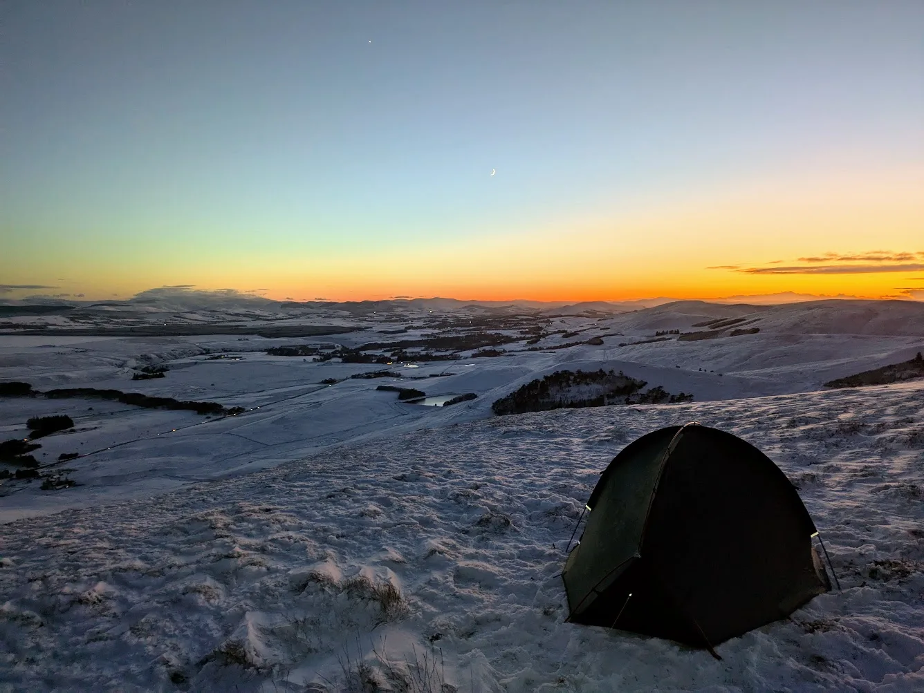 A dark backpacking tent pitched on a snowy hillside at sunset